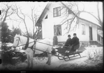 2 män sittande på hästekipage. Kvinna stående framför fönster på byggnad.
Nyckelord: Häst Släde Vinter