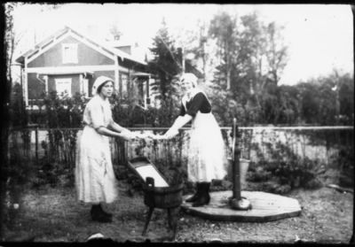 2 kvinnor stående och tvättar vid vattenpump.
Nyckelord: Pump Tvättbräda Hink