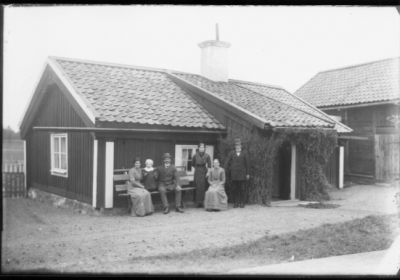 2 män, 3 kvinnor och ett barn sittande och stående vid bänk framför byggnad.
Nyckelord: Bänk