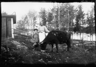 Kvinna och ett barn stående med en ko framför uthus.
Nyckelord: Ko