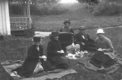 Man och 4 kvinnor sittande och drickandes kaffe på backen.
Nyckelord: Kafferast Picnic Filt Dragspel Dräkt
