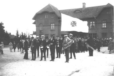 Ett antal personer stående framför byggnad.
Nyckelord: Flagga Musikinstrument