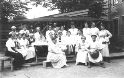 Ett antal personer sittande och stående framför byggnad.
