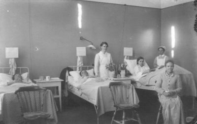 6 kvinnor, 3 liggande, 2 stående och 1 sittande i sjukhussal.
