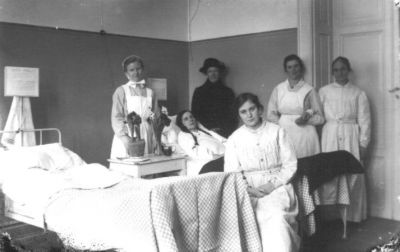 6 kvinnor, 1 liggande, 4 stående och 1 sittande i sjukhussal.
