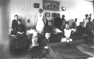 2 stående kvinnliga sköterskor och ett antal barn sittande, stående och liggande vid sängar i sjuksal.
