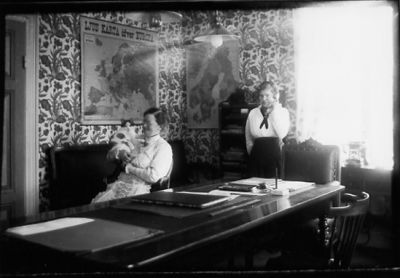 Kvinna sittande med katt i knät och kvinna stående bakom.
Nyckelord: Katt Skrivbord Telefon Väggkarta