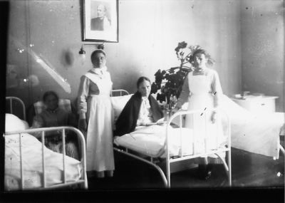 Kvinna sittande i säng och 2 kvinnor stående bredvid.
Nyckelord: Säng Uniform