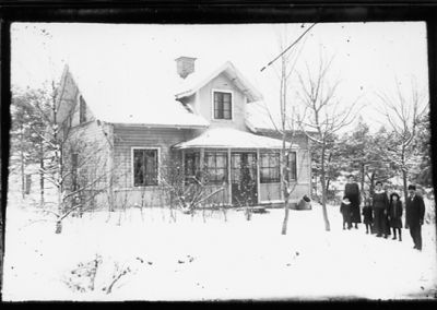 Familj stående i trädgård framför byggnad.
Nyckelord: Vinter