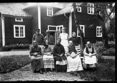 2 stående män, 2 stående kvinnor och 5 sittande kvinnor framför byggnad.
Nyckelord: Parkbänk Bostad