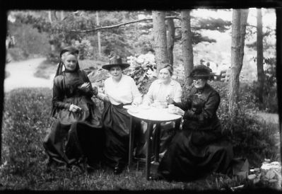 4 kvinnor sittande vid kaffebord.
Nyckelord: Trädgårdsmöbel Skog Dräkt Hatt