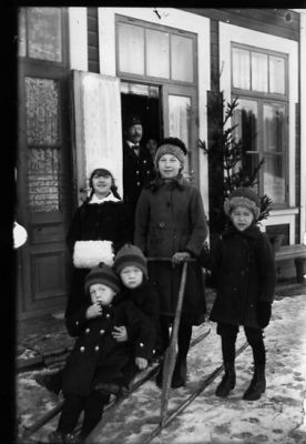 3 barn stående vid och 2 barn sittande på spark. Man och kvinna stående i dörröppning.
Nyckelord: Spark Muff Vinter