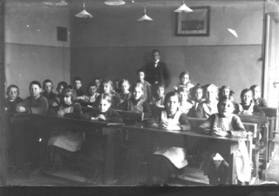 Skolklass sittande i klassrum med magister stående längst bak.
