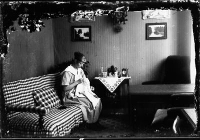 Kvinna sittande i soffa i rum med handarbete i knät.
Nyckelord: Tavla Handarbete