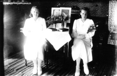 2 flickor sittande i rum med blommor i famnen och på bord.
Nyckelord: Blommor