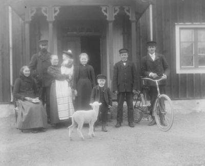 6 vuxna 2 barn och 1 lamm framför byggnad
Nyckelord: Cykel Bok Lamm Dräkt
