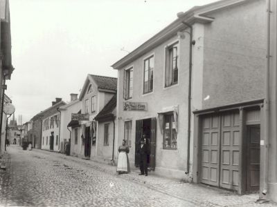 Gatuvy med 3 personer stående vid entren till tobaksaffär
Nyckelord: Affärsskylt Dräkt Kullersten