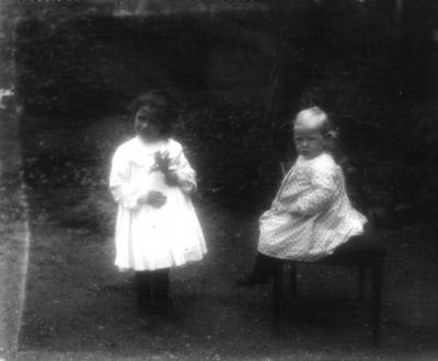 1 stående och 1 sittande flicka i trädgård
Nyckelord: Trädgård
