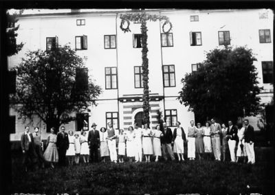 Ett antal personer stående framför midsommarstång och byggnad.
Keywords: Midsommarstång