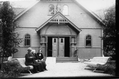 Man och 2 kvinnor sittande på bänk framför kyrka.
Nyckelord: Parkbänk