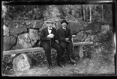 2 män sittande på bänk intill vattendrag.
Nyckelord: Käpp Bänk