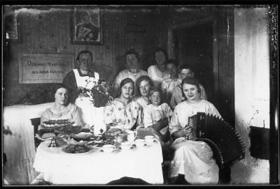 4 kvinnor och 1 pojke stående och 4 kvinnor och 1 pojke sittande vid kaffebord i rum.
Nyckelord: Kaffe Dragspel Väggbonad