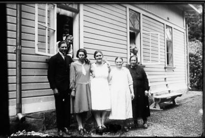 Man och 4 kvinnor stående framför byggnad och 4 personer i fönstren.
