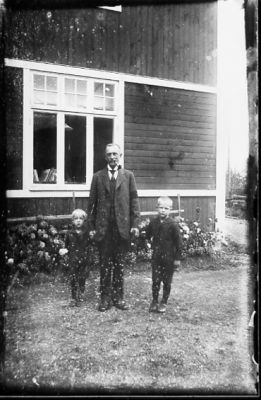 Man och 2 pojkar stående framför byggnad.
