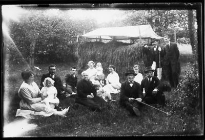 Ett antal personer stående eller sittande framför hässja i hage.
Nyckelord: Hässja