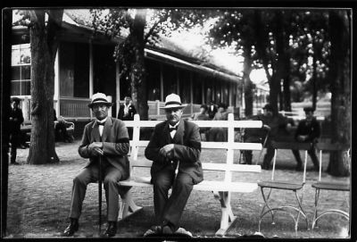 2 män med sorgband sittande på parkbänk med byggnad i bakgrunden.
Nyckelord: Parkbänk Käpp Sorgband