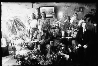 Man och 4 kvinnor stående och 2 män sittande i rum med blommor.
Nyckelord: Blommor Tavla