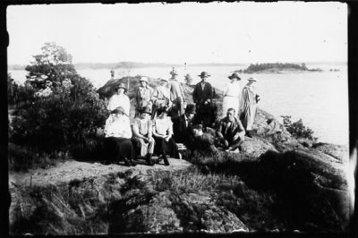 Ett antal personer stående eller sittande på berg med vatten och öar i bakgrunden.
Nyckelord: Ö