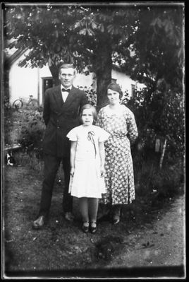 Man, kvinna och flicka stående i trädgård.
