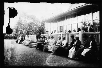 Ett antal personer sittande på och nedanför veranda.
Nyckelord: Veranda