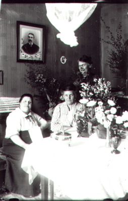 2 kvinnor sittande 1 stående vid bord i rum.
Nyckelord: Blommor Tavla
