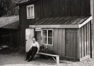 Man sittande på bänk framför låg träbyggnad
Nyckelord: Hus Skomakeri