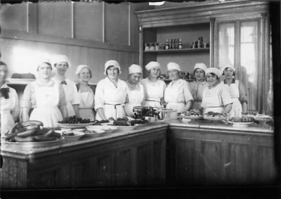 11 kvinnor stående bakom disk i kök.
Nyckelord: Dräkt