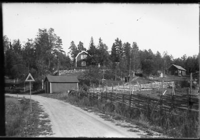 Landsväg med gärdesgård vid sidan och byggnader i bakgrunden.
Nyckelord: Gärdesgård