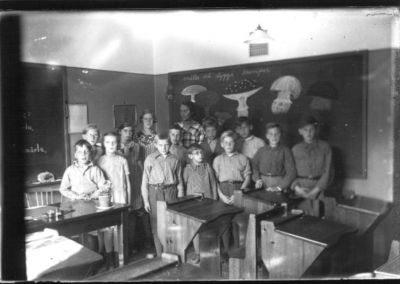 13 skolbarn stående i klassrum med skolfröken längst bak.
Nyckelord: Skolsal Svarta tavlan