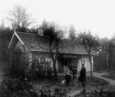 Man, kvinna, pojke och hund stående i trädgård framför byggnad.
Nyckelord: Hund