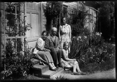 Man, kvinna och flicka sittande, flicka stående på trappa till byggnad.
Nyckelord: Trädgård