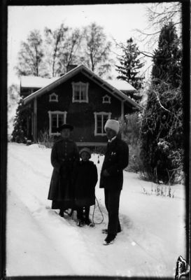 Man, kvinna och flicka stående i snön framför byggnad.
Nyckelord: Vinter