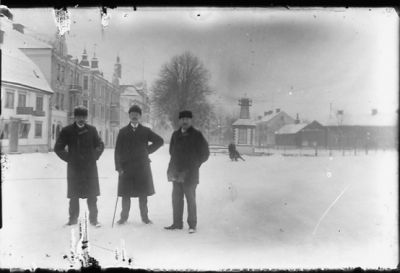 3 män stående i snön på torg med byggnader i bakgrunden.
Nyckelord: Käpp Vinter