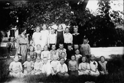Ett antal barn och 1 kvinna stående eller sittande på gräsmatta.
Nyckelord: Skolklass Gräsmatta