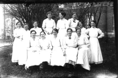 8 kvinnor stående och 5 kvinnor sittande framför byggnad.
Nyckelord: Park Byggnad Lasarett Uniform