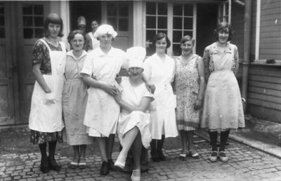 8 kvinnor stående och 1 sittande framför byggnad.
Nyckelord: Gatsten Fönster Byggnad