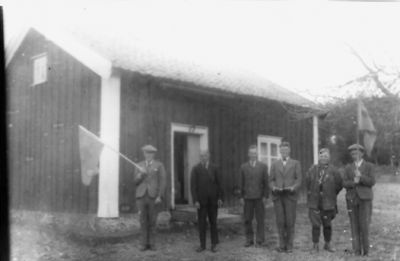 6 män stående framför byggnad. 2 med flagga i hand.
