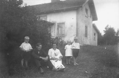 Man, kvinna och 4 barn framför byggnad.
Nyckelord: Bostad