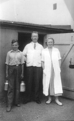 Man och kvinna stående i dörröppning och pojke stående med mjölkkrukor i händerna.
Nyckelord: Mjölkkruka Dörröppning