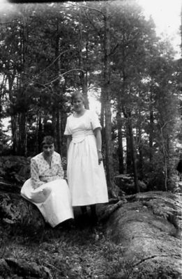 Kvinna stående och kvinna sittande i skogsbacke.
Nyckelord: Skogsbacke Skog